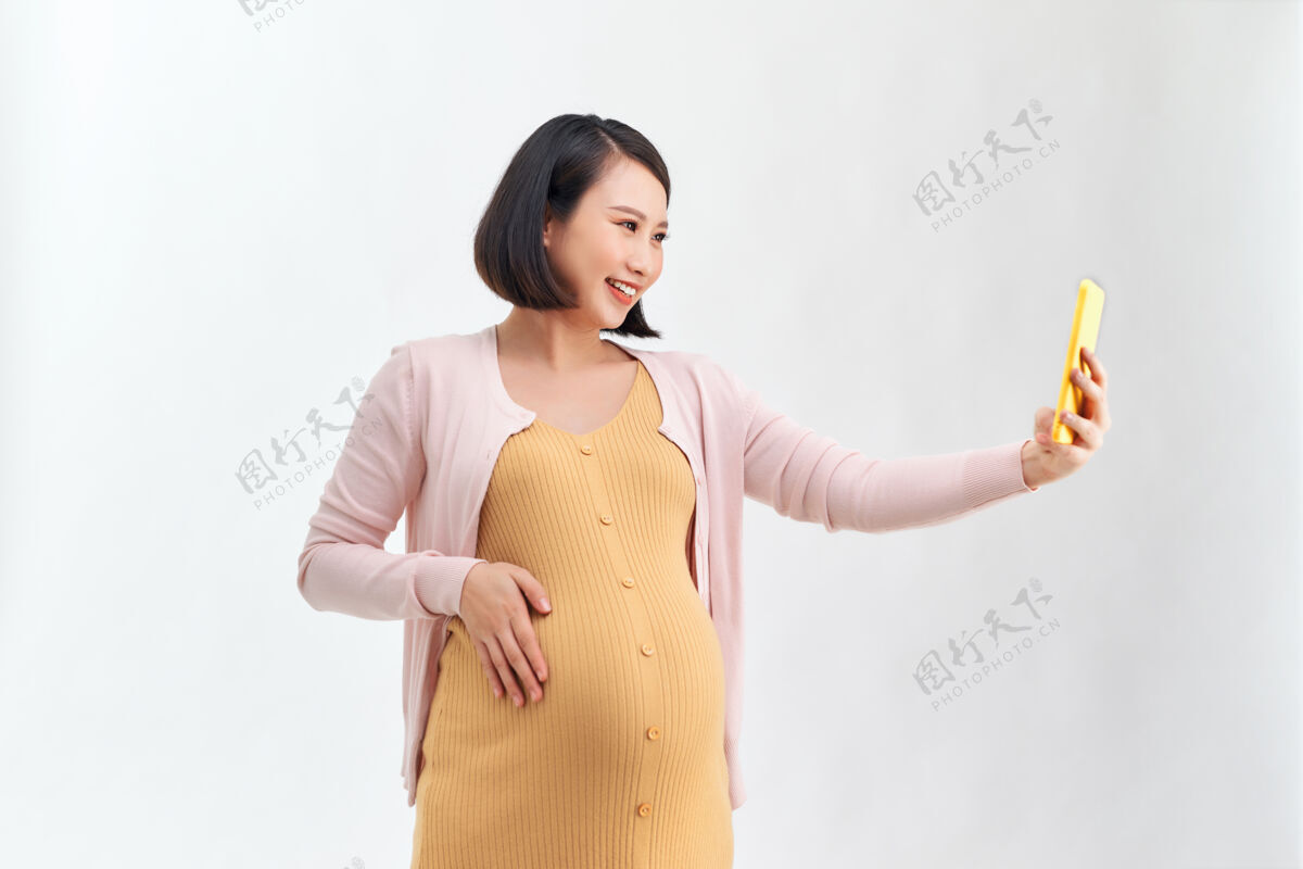 漂亮快乐的孕妇与智能手机采取自拍超过白色亚洲人成年人肚子