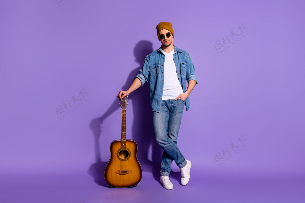 生活方式全身尺寸的照片 自信的吉他倚靠在他的吉他上戴着帽子白色运动鞋鞋隔离在紫色鲜艳的颜色背景投下阴影休闲吉他手男人