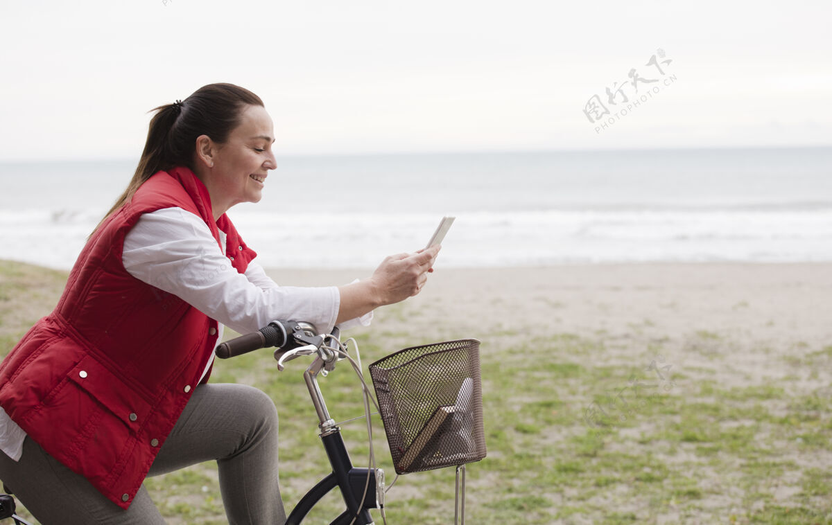 微笑迷人的成熟女子骑自行车使用手机海滩背景生活方式快乐互联网