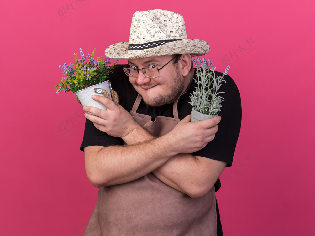 人贪婪的年轻男园丁戴着园艺帽 拿着和粉色墙壁隔离的花盆里的花在一起持有交叉帽子