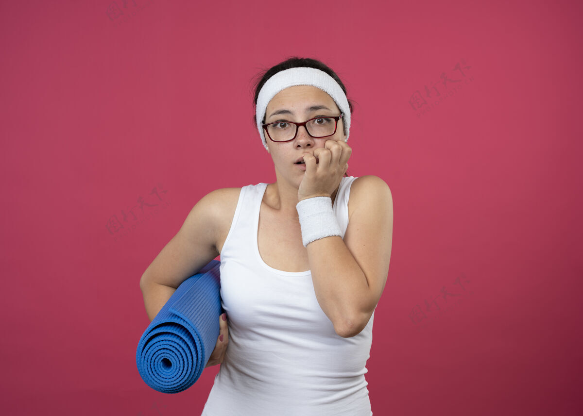 穿戴着眼镜 戴着头带和腕带 焦虑不安的年轻运动女性咬着指甲 把运动垫孤立地放在粉红色的墙上年轻垫子腕带
