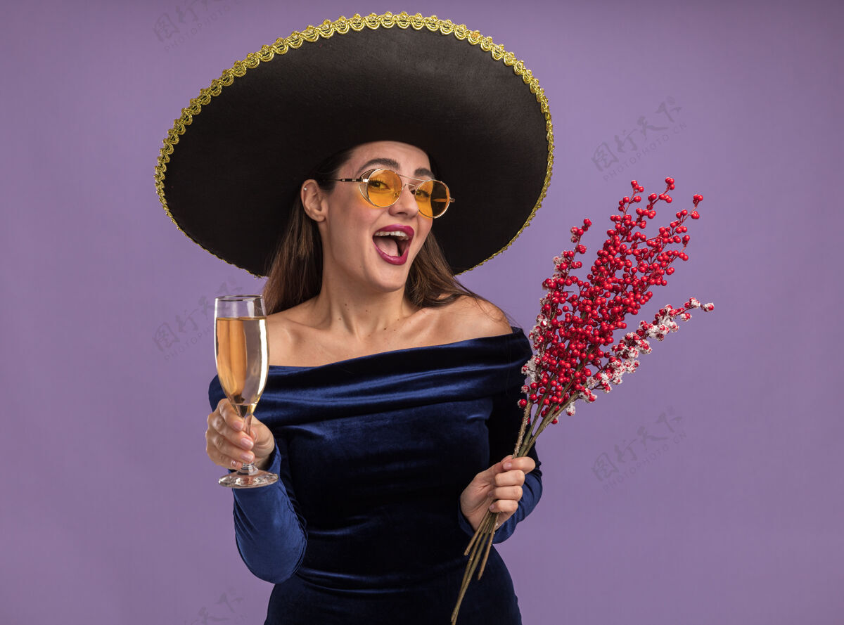 玻璃兴奋的年轻漂亮女孩穿着蓝色连衣裙 戴着墨镜 拿着罗文的树枝 在紫色的背景上放着一杯香槟罗文年轻连衣裙