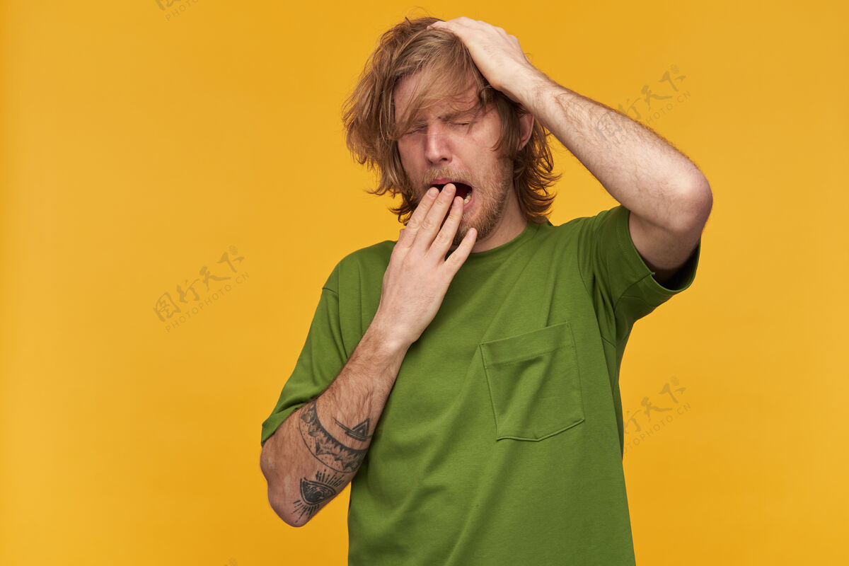 服装昏昏欲睡的男人 疲惫的男人 金色凌乱的头发 胡子和小胡子穿着绿色t恤有纹身抓他的头打哈欠遮住他的嘴站在黄色的墙上孤立年轻帅气金发