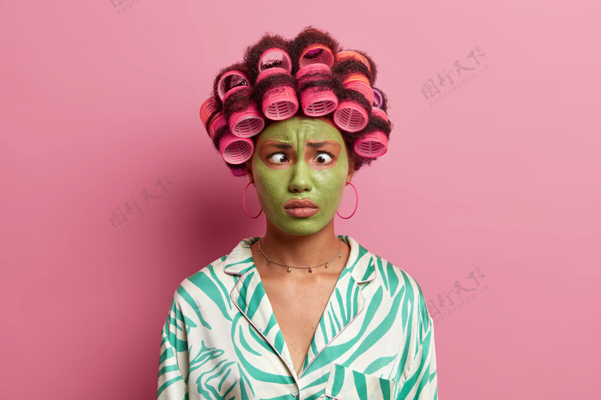 风格搞笑的民族女青年做鬼脸 交叉眼睛 涂发卷 为特殊的日子做发型 脸上戴绿色保湿面膜民族绿色面具