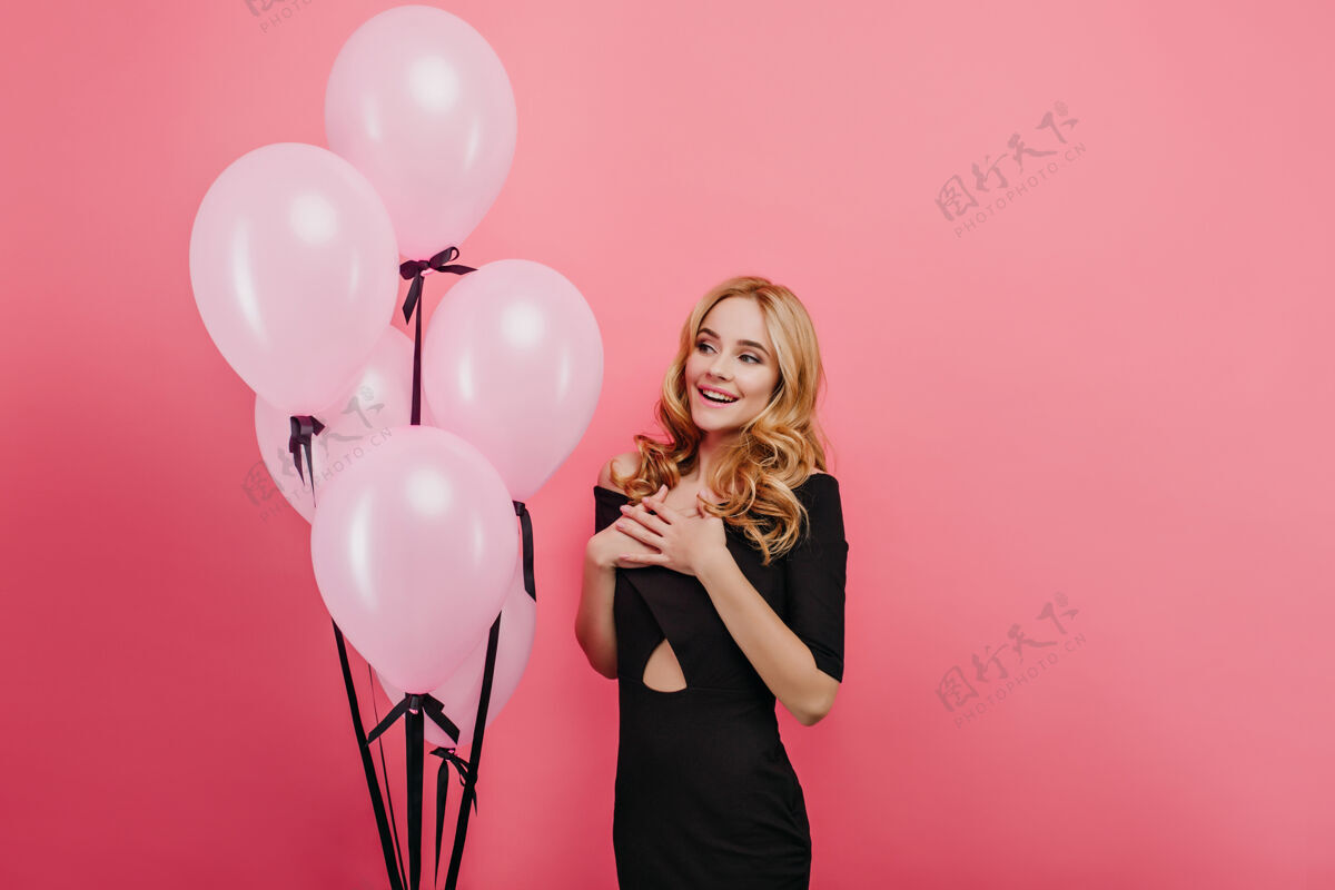 肖像美丽的卷发女人 化着时髦的妆容 在生日派对上露出惊讶的微笑苗条的金发女孩站在明亮的墙上的氦粉色气球旁情感表情成人