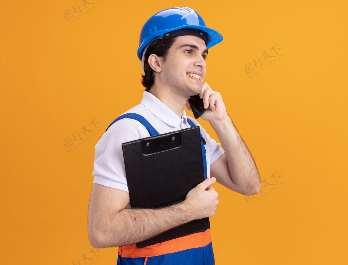 年轻人身穿施工制服 头戴安全帽的年轻建筑工人站在橙色的墙上 手里拿着剪贴板 微笑着讲着手机头盔建筑工人建筑