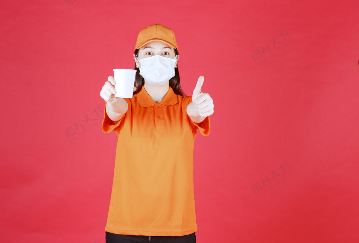 制服身着橙色制服和口罩的女性服务人员手持一次性杯子 并显示积极的手势新常态姿势面膜