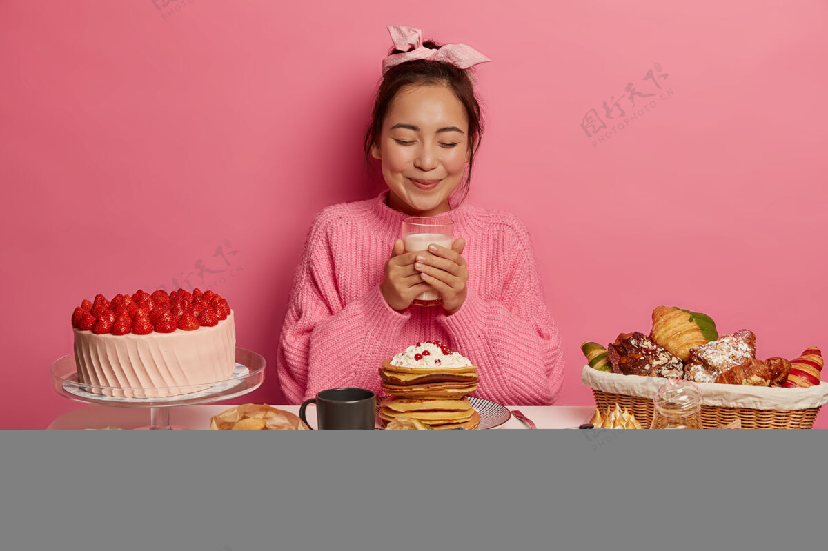 食物漂亮的黑发女人端着一杯牛奶 吃着糖果和糖果 穿着针织毛衣和头带 在粉色背景下的喜庆餐桌上摆着甜食的姿势饮料烘焙节日