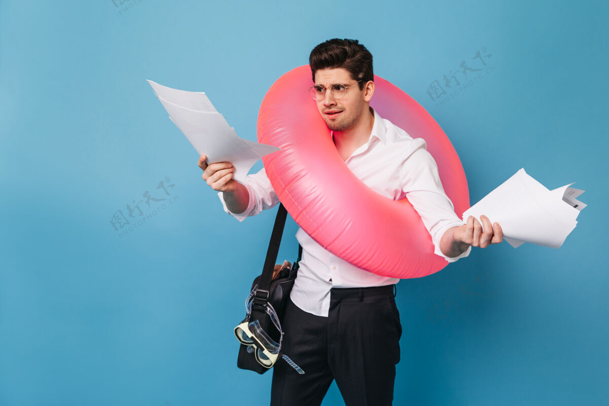 男性身穿办公服的黑发男人不高兴地看着文件这家伙要去旅行 摆出粉红色充气圈的姿势西装商人优雅