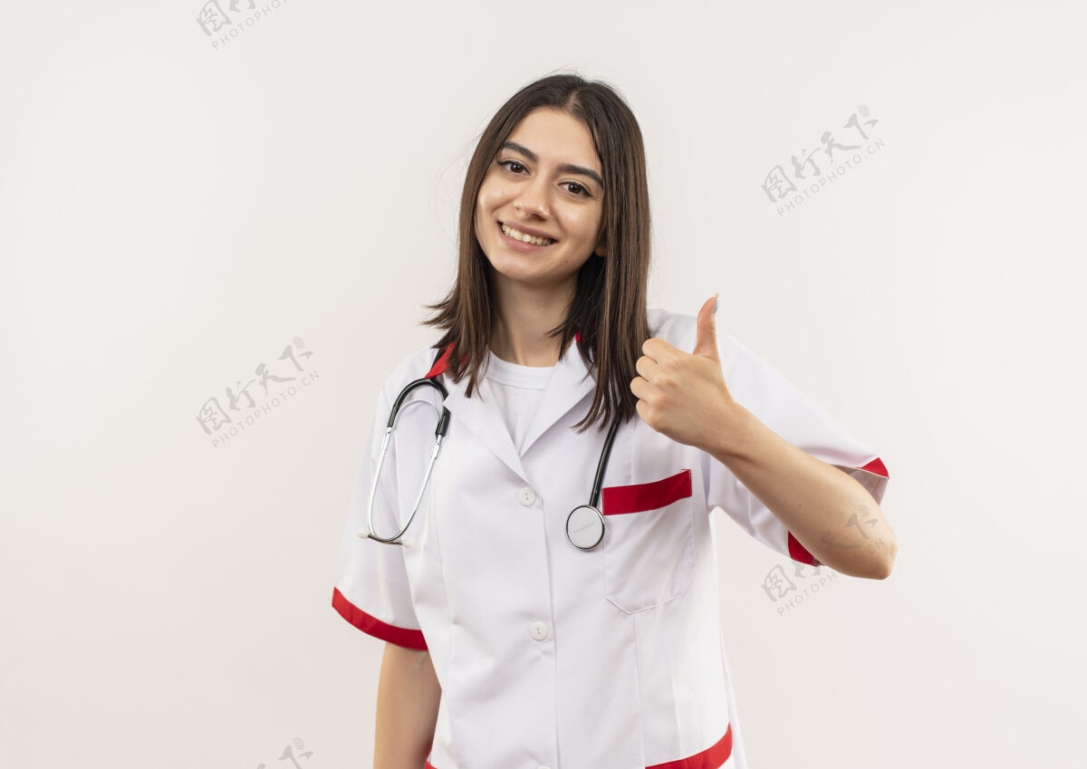 年轻人年轻的女医生穿着白大褂 脖子上戴着听诊器 面带微笑地站在白墙上竖起大拇指 望着前方周围女人听诊器