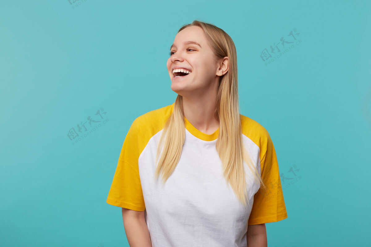 女性快乐的年轻迷人的长发金发女士 随意的发型 站在蓝色背景下 穿着白色和黄色的t恤 面带微笑 愉快地看着一边表情快乐肖像