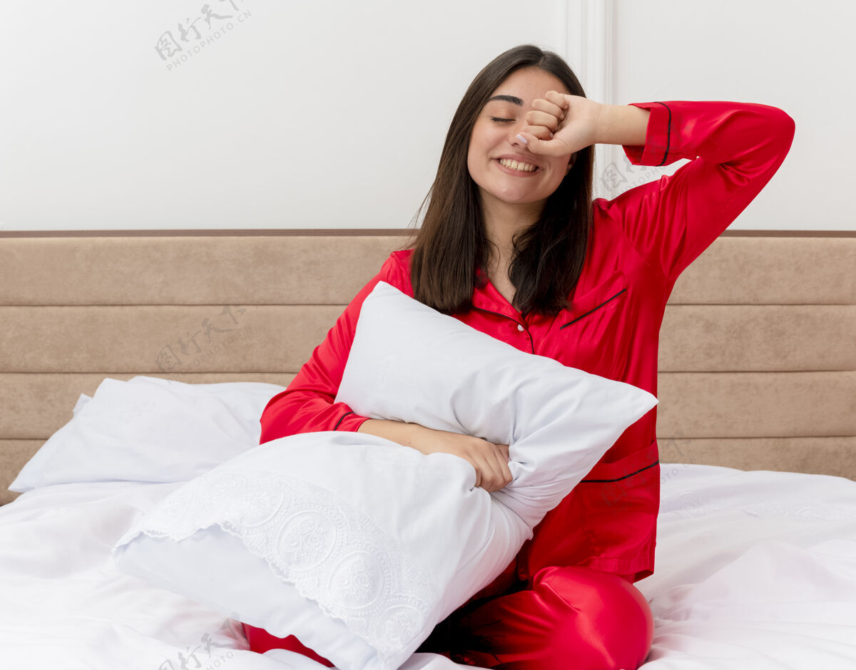 红色穿着红色睡衣的年轻漂亮女人坐在带枕头的床上 想在卧室里打呵欠睡觉 背景光线很浅床年轻坐着