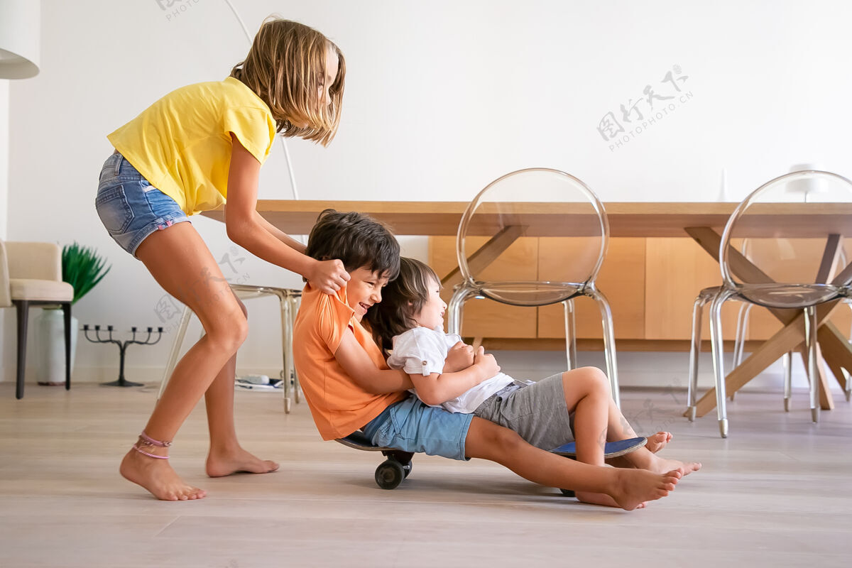 可爱快乐的孩子们在家玩滑板金发可爱的女孩推着她的两个顽皮的兄弟快乐的孩子们骑在滑板上玩童年 游戏活动和周末的概念白种人年轻室内