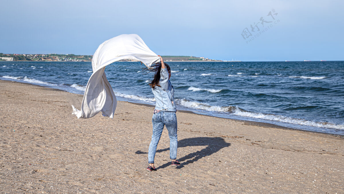 风海边的年轻女子在风中抱着一张大张床单 享受着自由的生活方式 度假自由女人