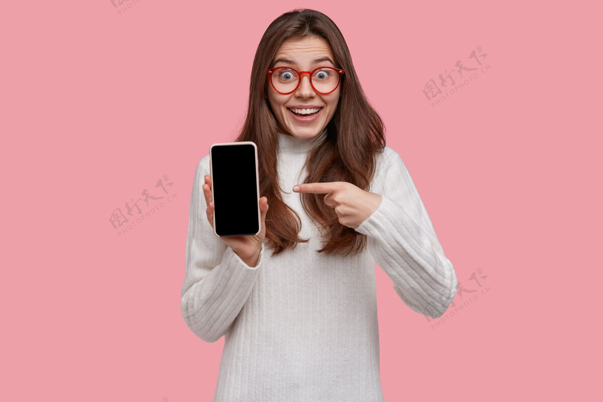 保持激动得说不出话来的年轻女子指着智能手机模拟屏幕 试图展示一些令人敬畏的东西 穿着白色套头衫手机指向情绪