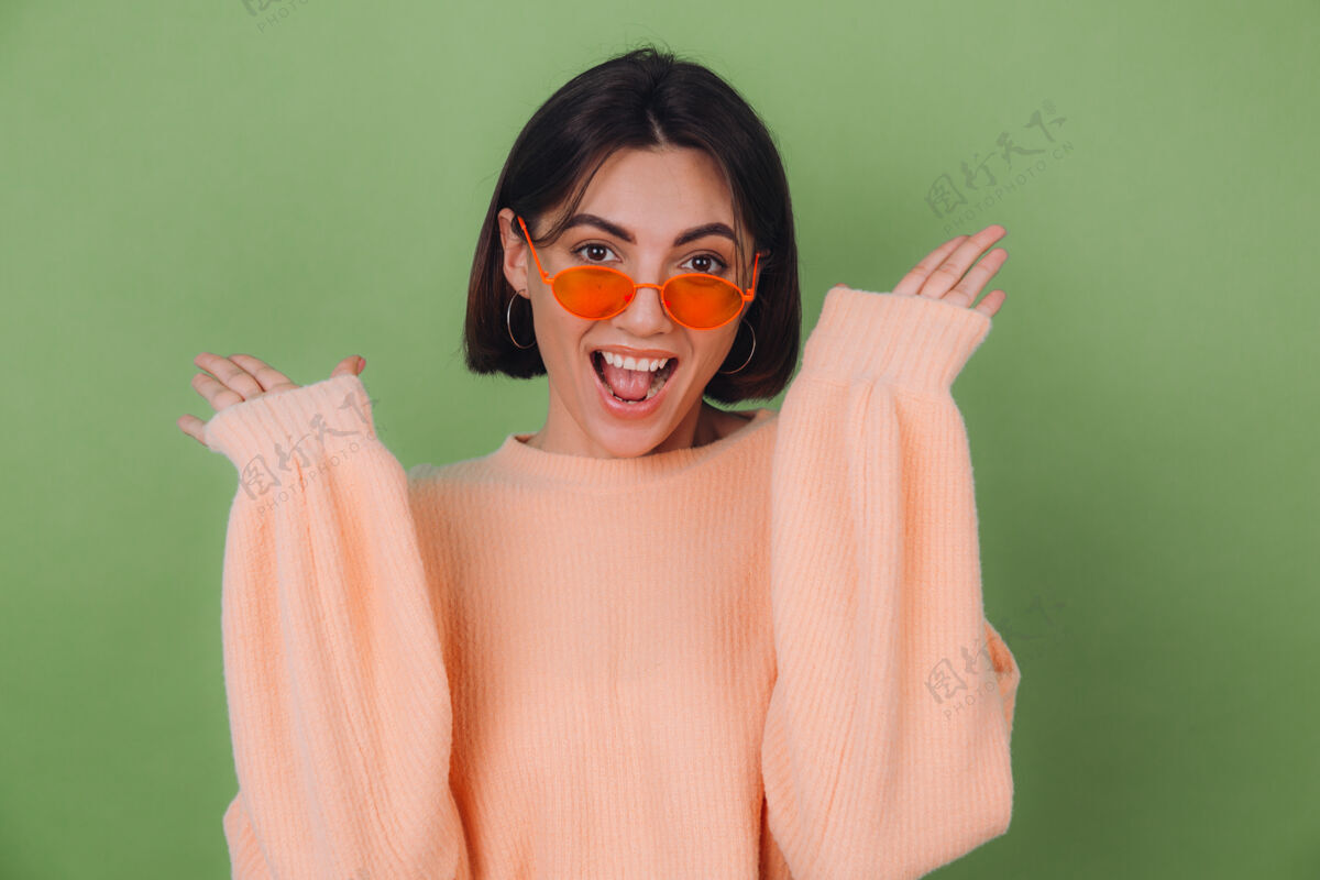 人穿着休闲桃色和橙色眼镜毛衣的年轻女子隔离在绿橄榄墙上兴奋地张嘴摊开双手积极时尚惊人