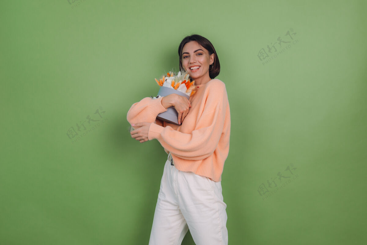 女性年轻女子穿着休闲桃色毛衣隔离在绿橄榄墙上手持橙白色花盒组成的棉花 吉普赛拉小麦和拉古鲁斯作为礼物开心惊喜静物帽盒内部