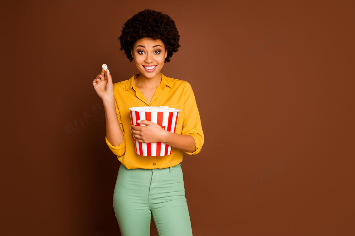 年轻搞笑迷人的深色皮肤卷曲女士拿着爆米花桶吃玉米看最喜欢的电视节目穿黄色衬衫绿色裤子孤立棕色的照片女孩多国多民族