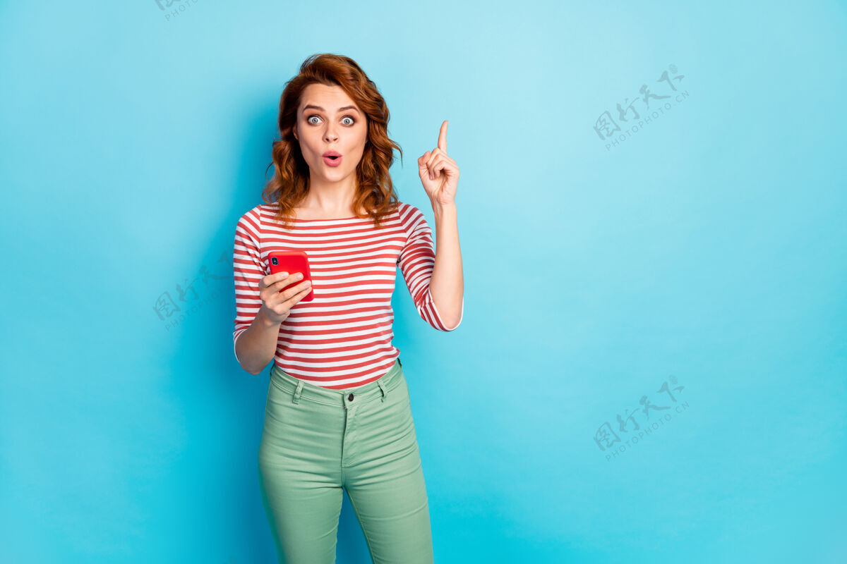裤子惊讶女人的画像用手机聊天发帖思考有问题有答案举起食指印象深刻尖叫哇天哪穿毛衣孤立的蓝色红发漂亮移动