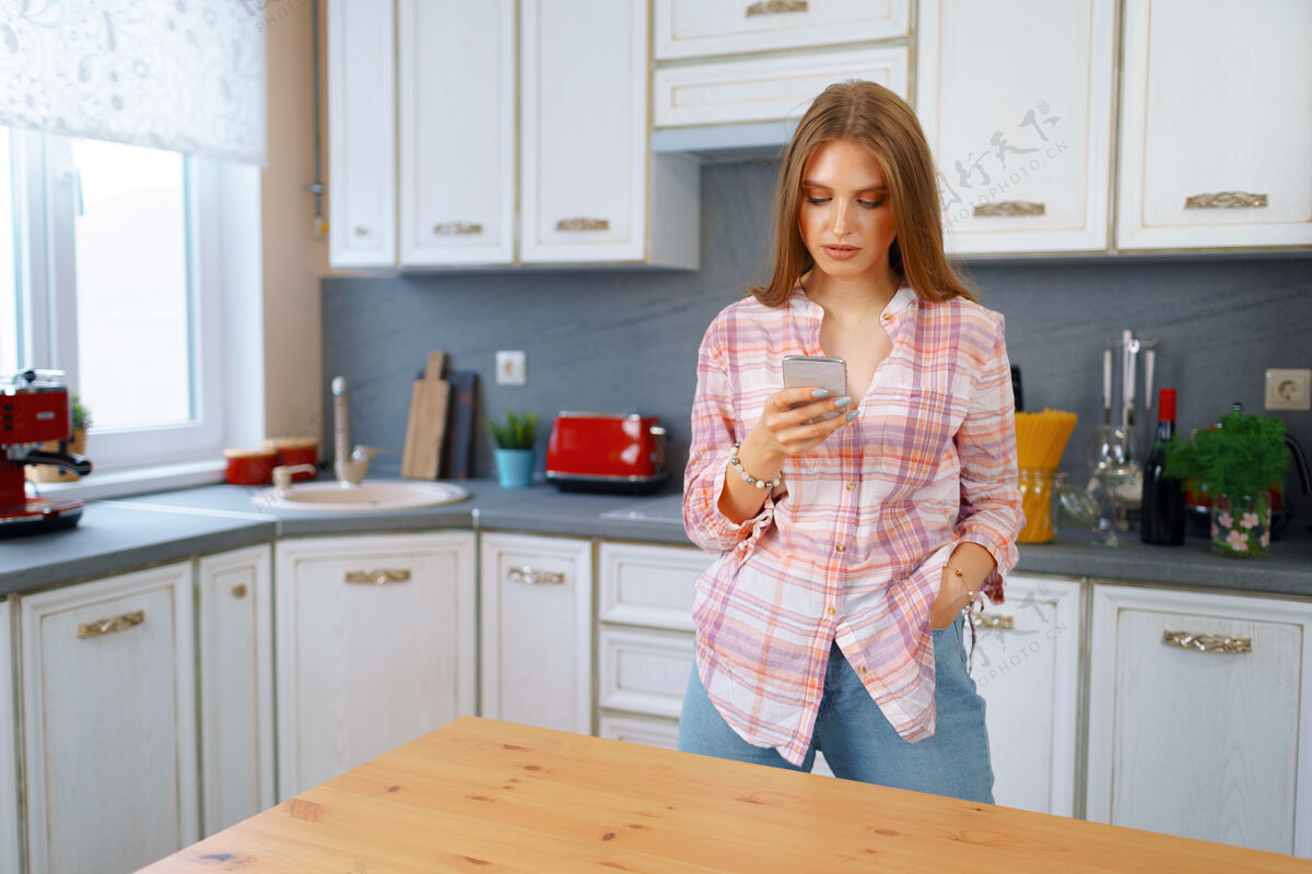 通讯穿着休闲服的年轻漂亮女人在厨房里用她的智能手机女孩室内白种人