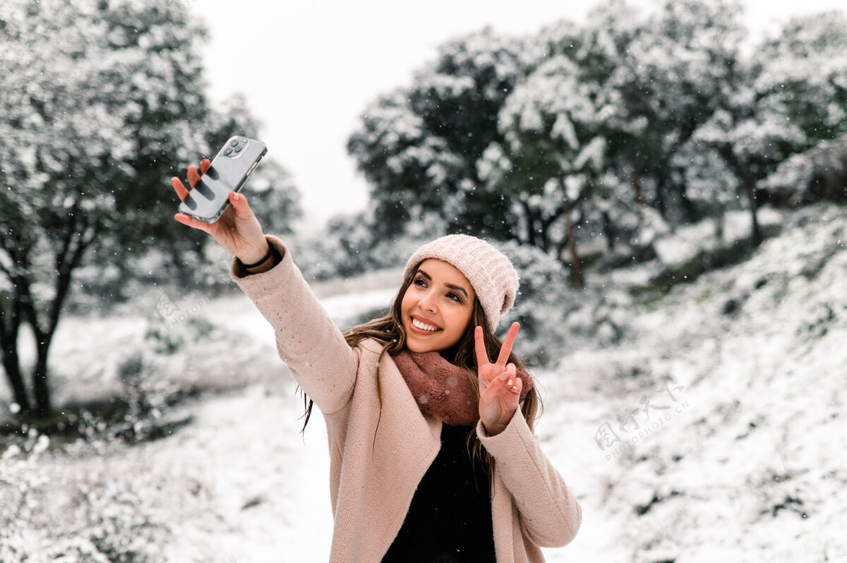 阴天穿着暖和外套的女人站在树林里 一边用智能手机自拍雪景外套风景寒冷