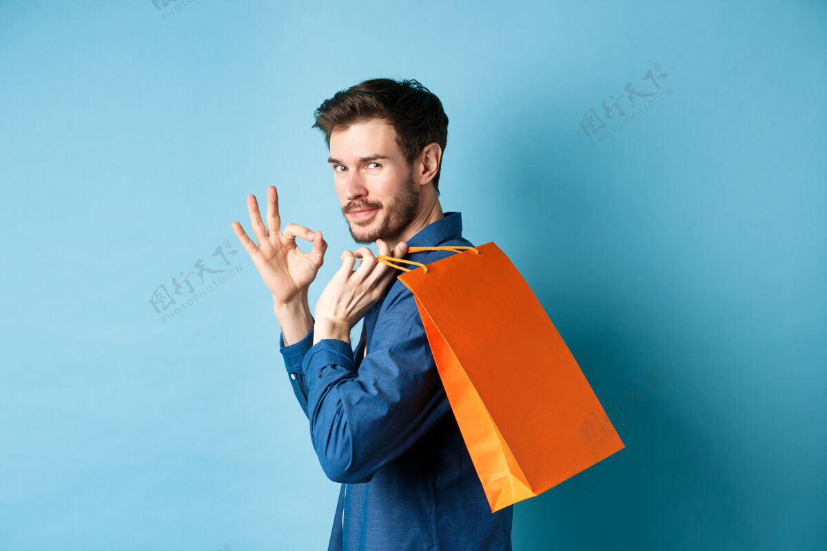 包帅哥肩上扛着购物袋 转过身来看着镜头 摆出OK的姿势 推荐店铺 蓝色背景男性人买家