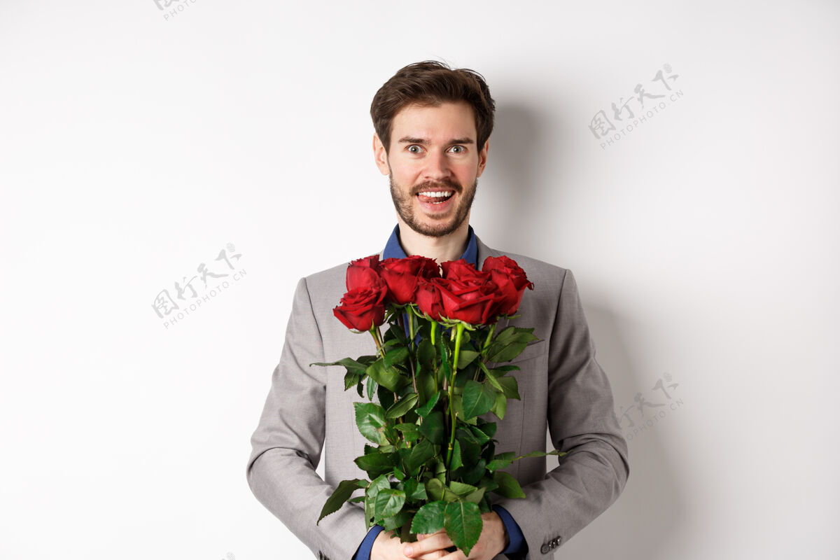 年轻穿着西装的兴奋帅哥捧着玫瑰花束与情人浪漫约会 在情人节快乐地站着 白色背景帅气浪漫情人