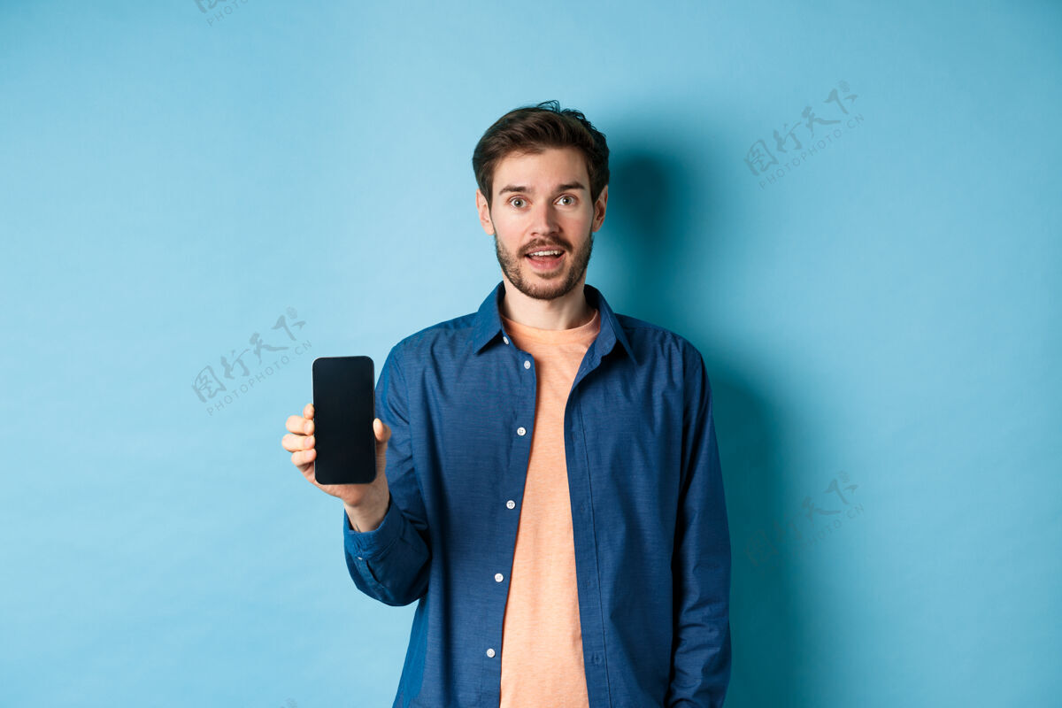 移动快乐的白种人站在蓝色的背景上 展示着空荡荡的手机屏幕和微笑社交电话表情