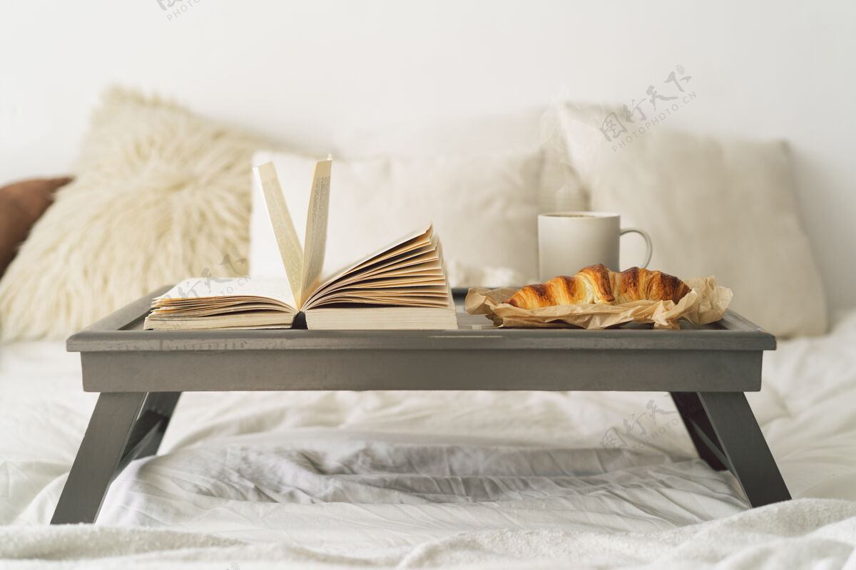 家具托盘上放着牛角面包 一杯咖啡和一本书 放在白色干净的床上床阅读杯子