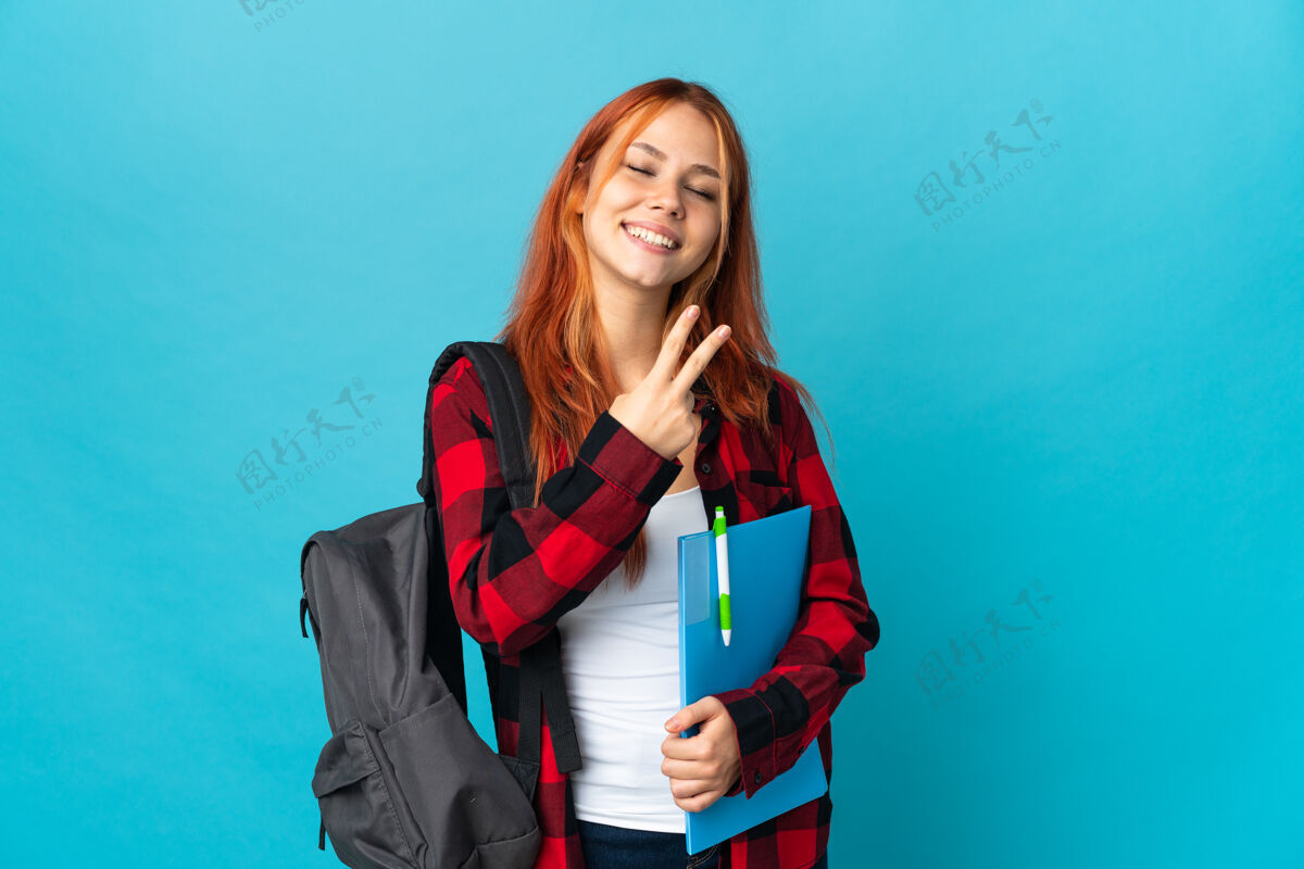 大学学生俄罗斯女子孤立在蓝色微笑和显示胜利的标志乐趣手势女人