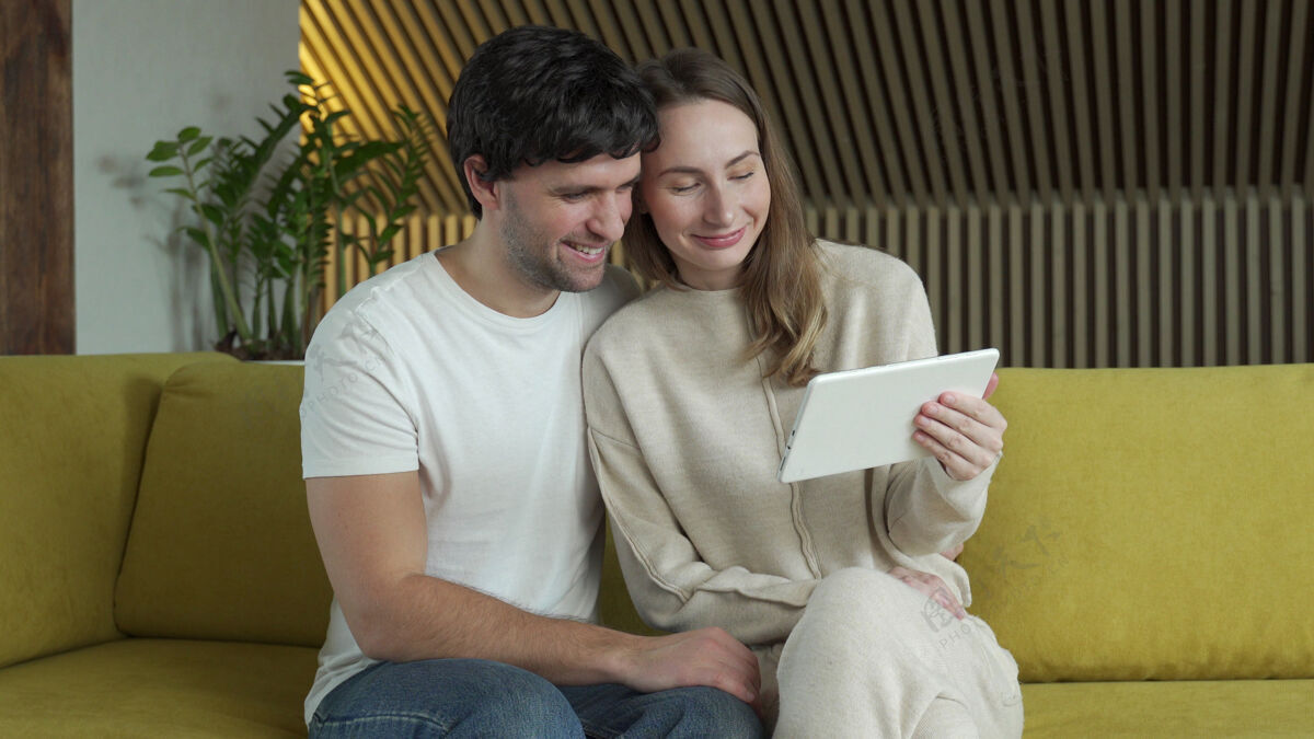沙发坐在家里沙发上 坐在平板电脑里看网络媒体内容的年轻夫妇情人Wifi笑