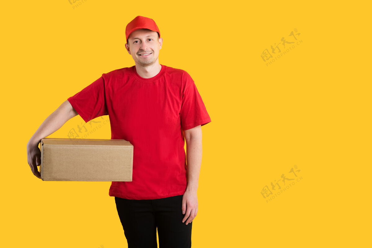 微笑快乐的年轻送货员穿着红色制服站在黄色的纸箱上包装黄色信使