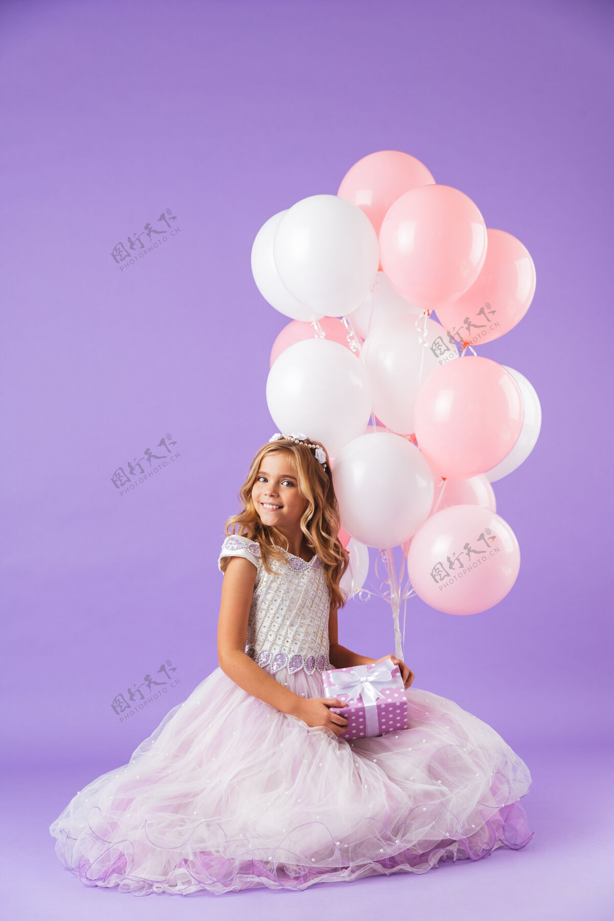 欢乐穿着公主裙的漂亮小女孩孤零零地坐在紫罗兰色的墙上 手里拿着一堆气球和一个礼盒微笑白种人气球