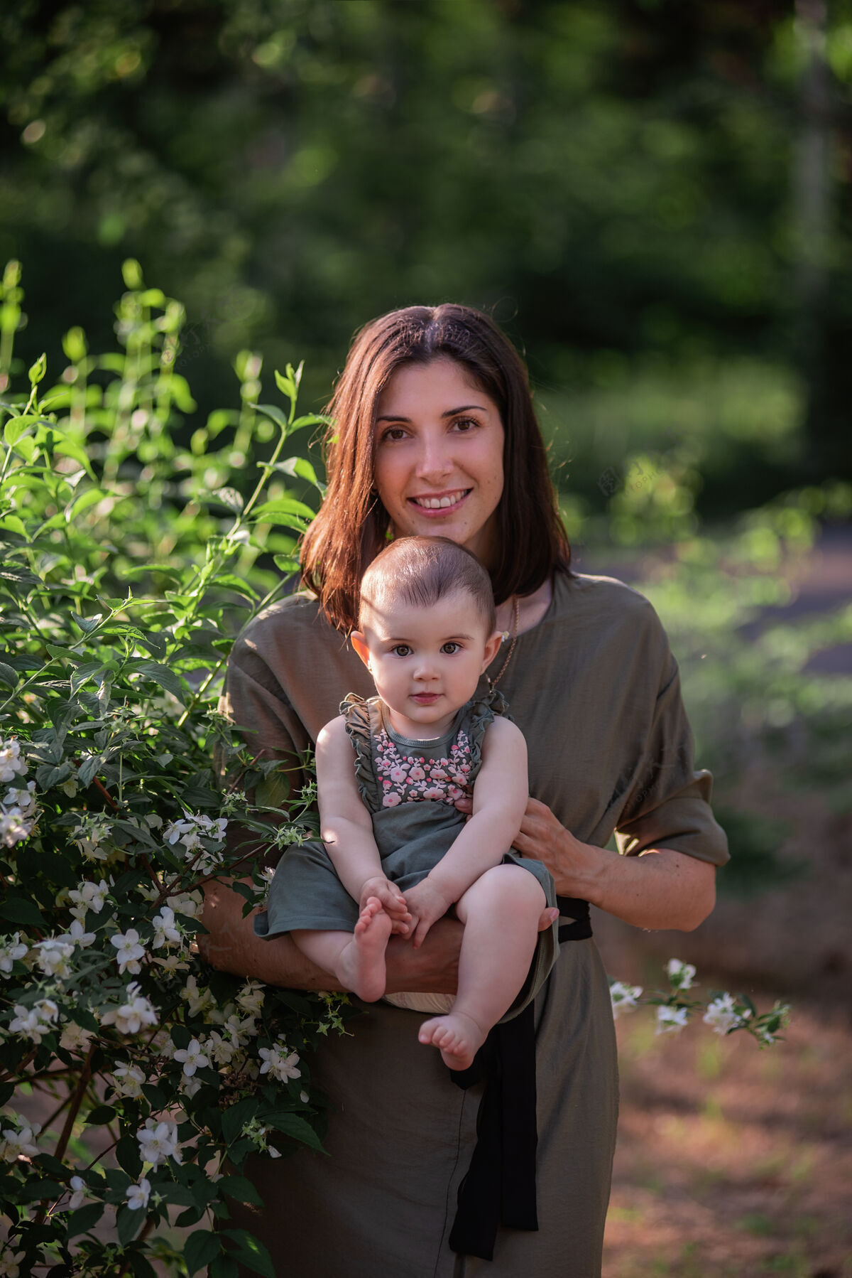 茉莉花一个年轻的女人怀里抱着一个小婴儿一位美丽的母亲和她的女儿在茉莉花丛旁的绿色公园里散步可爱小草宝宝