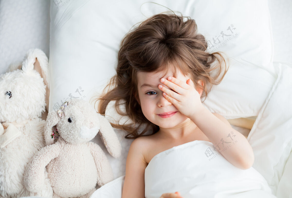 梦可爱的小女孩把她的眼睛藏在她的手后面醒来了她他和兔子玩具躺在白色的床上微笑甜蜜童年