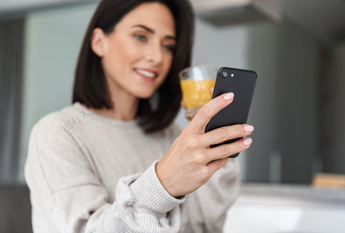 女士满意的女人30多岁喝橙汁和使用手机 而在明亮的现代房间休息饮料吸引力舒适