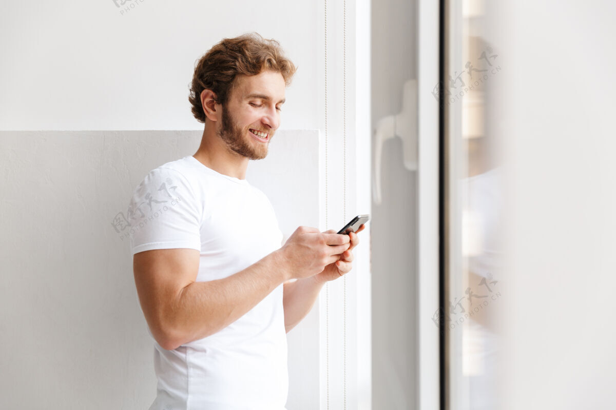 成人微笑的年轻人拿着手机 站在家里的窗前肖像交谈吸引力