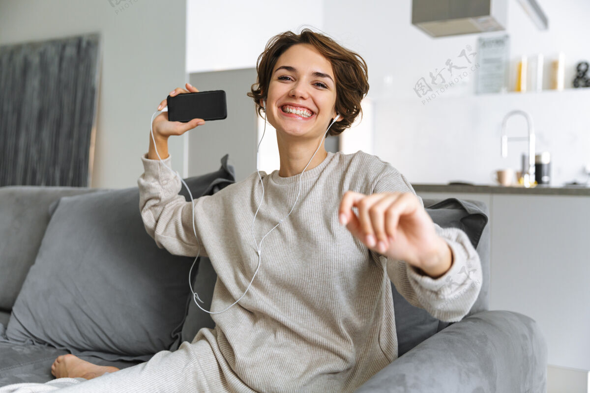 娱乐快乐的年轻女子坐在家里的沙发上 用手机 用耳机听音乐音乐沙发声音