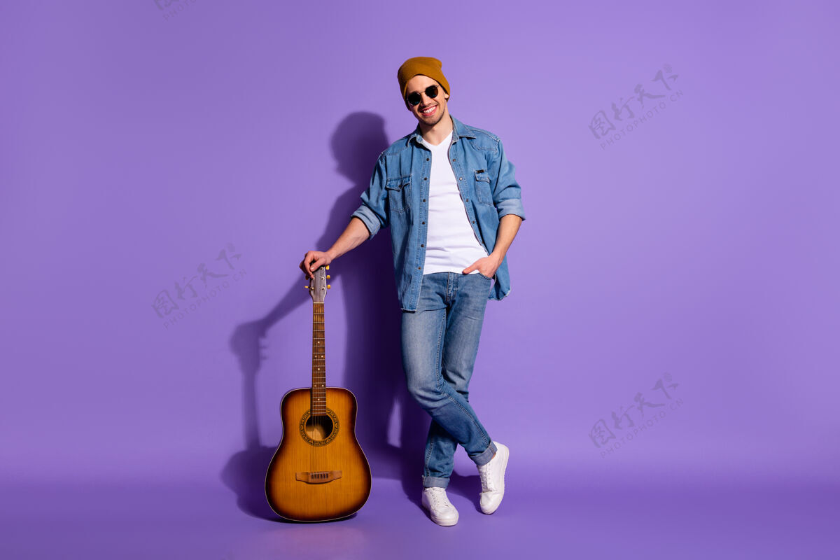 吉他手全身尺寸照片欢快积极可爱帅气的音乐制作人靠在他的吉他上穿着帽子运动鞋鞋鞋孤立的紫色明亮的颜色背景欢乐积极爱好