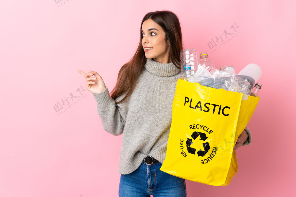 看年轻的白种女人拿着一个装满塑料瓶的袋子 在粉红色的背景上用手指着一边回收指向介绍重复使用