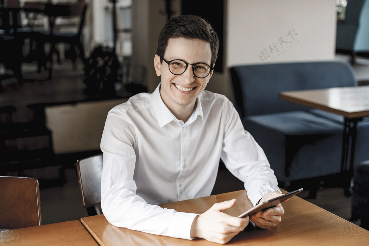 房子一个积极英俊的年轻男性坐在书桌旁 面带微笑地看着相机 手里拿着一个戴着眼镜的平板电脑站立男商务人士