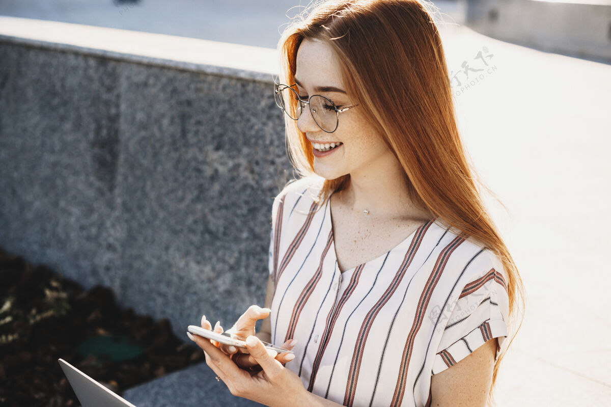 在线一位留着红发和雀斑的美丽年轻女子坐在一栋楼的楼梯上 闭着眼睛笑着 手里拿着一部智能手机年轻使用成人