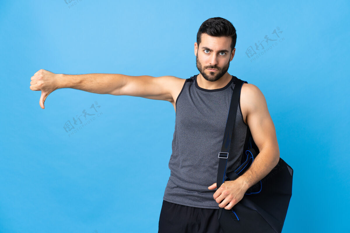 手指年轻的运动男子与运动包隔离在蓝色背景显示拇指向下与消极的表情站立健身房不喜欢