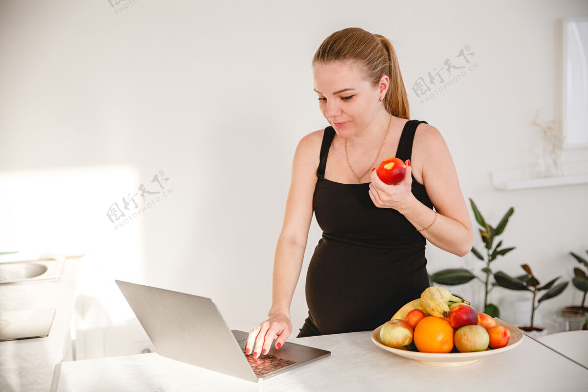 检疫年轻的金发孕妇穿着黑色连衣裙在白色厨房里吃水果 看着我笔记本电脑高高质量的照片女性情绪年轻