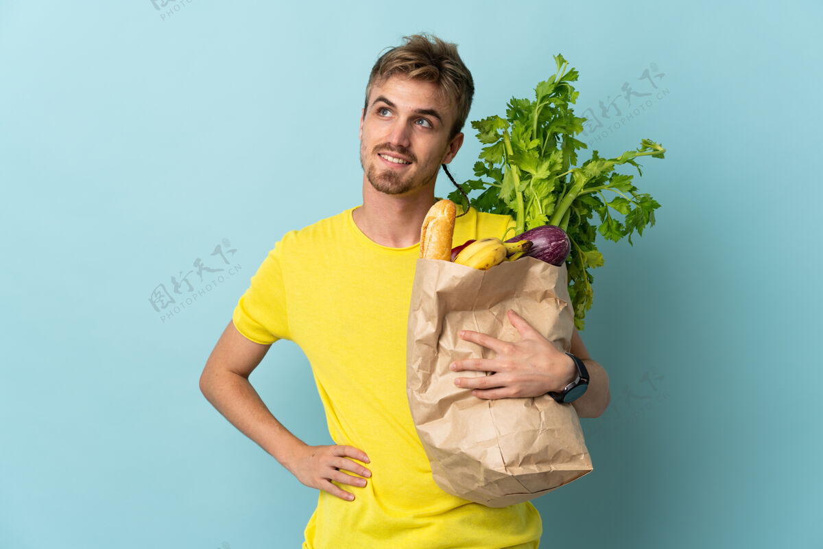 食物一个金发碧眼的人拿着一袋外卖食物孤立地站在蓝色的椅子上 双臂放在臀部 面带微笑素食营养帅哥