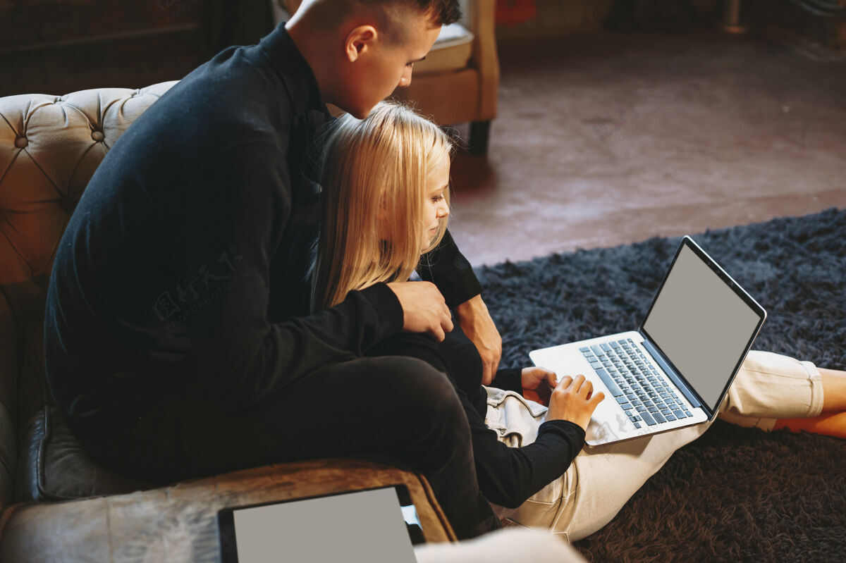金发漂亮的年轻女性在家里拿着笔记本电脑在地板上工作 而她的男朋友正从背后拥抱着她情侣年轻家庭