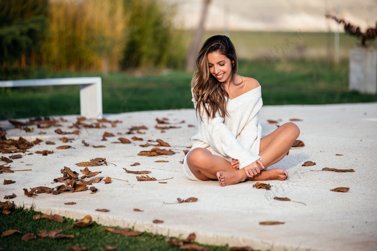 漂亮秋天 院子里的落叶旁 快乐的赤脚女人盘腿坐在水泥地上微笑着女孩微笑毛衣