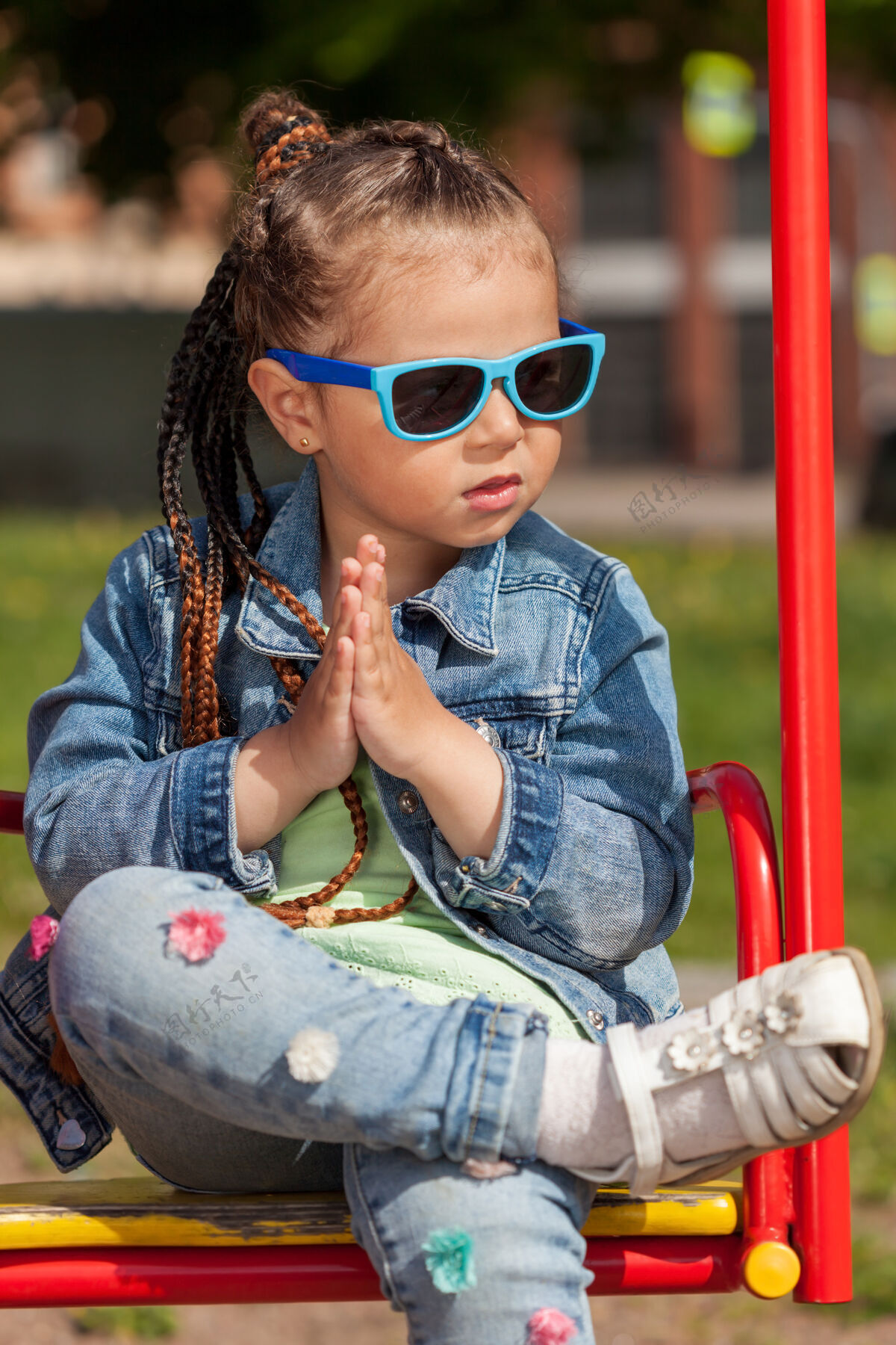 牛仔裤梳着辫子戴着太阳镜的时髦女婴坐在秋千上双手紧握牛仔黑发眼镜