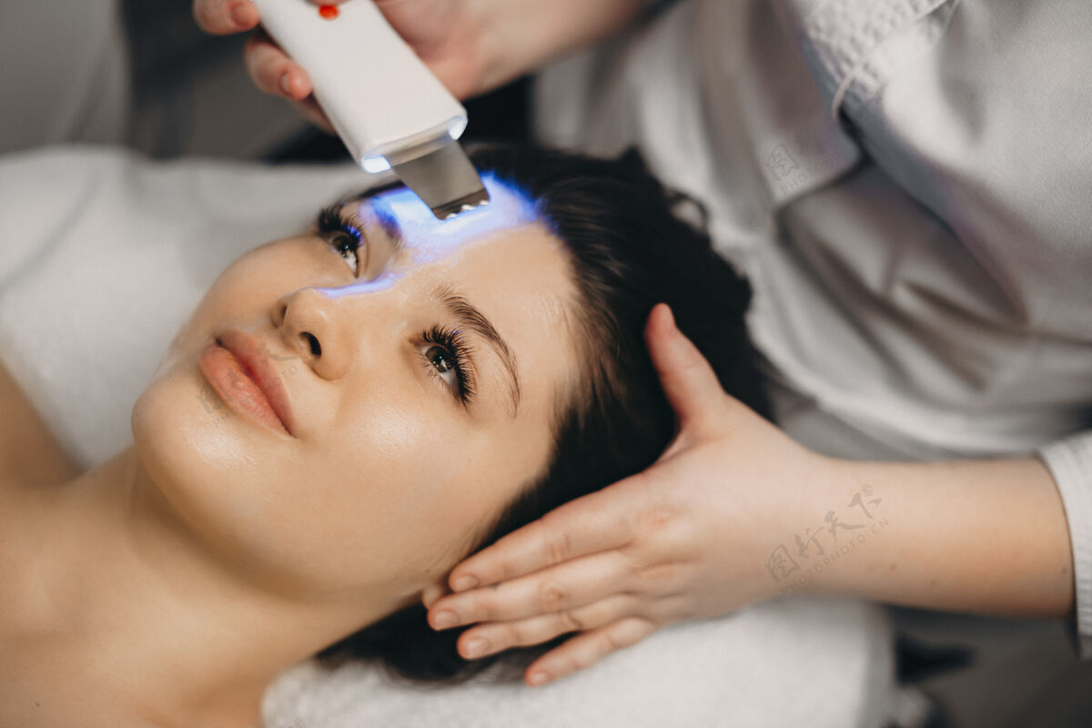 化妆品一位美丽的黑发女性躺在水疗床上睁着眼睛 同时由一位女性美容师对她的脸进行超声波检查特写皮肤护理人