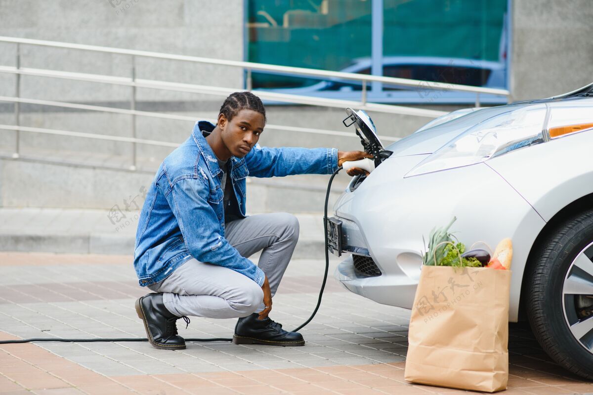 环境英俊的美国黑人男子坐在他的新的现代电动车旁边 拿着充电器的插头运输改变电力